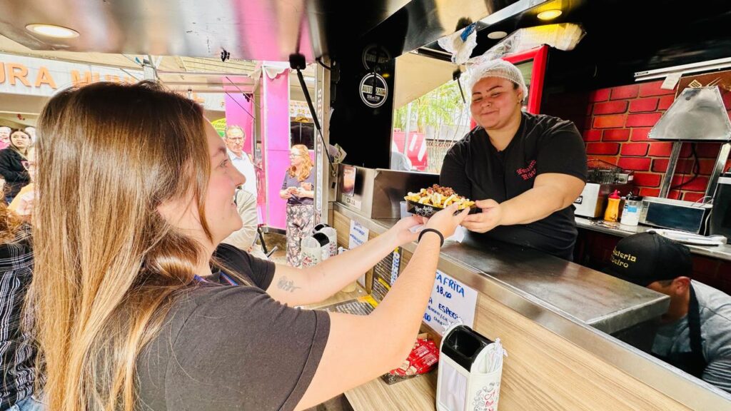 A imagem retrata uma cena vibrante em um food truck, onde uma funcionária, usando touca descartável, entrega um prato de batatas fritas com coberturas a uma cliente. A cliente, com cabelos castanhos longos, estende as mãos para receber o pedido. Ao fundo, outras pessoas aguardam, indicando um ambiente movimentado. O food truck possui uma decoração rústica, com tijolos aparentes e letreiros, sugerindo um local de comida informal e popular. A iluminação contribui para a atmosfera acolhedora e animada do local.