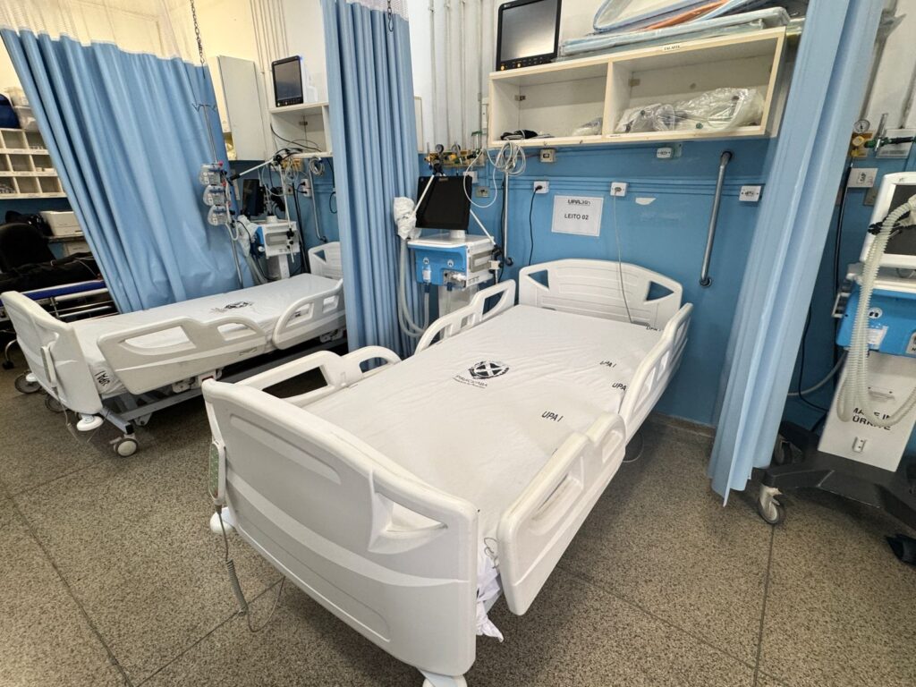 A imagem mostra um quarto de hospital com duas camas de hospital brancas e totalmente equipadas. As camas estão posicionadas lado a lado, com cortinas azuis separando-as e criando privacidade para cada paciente. Cada cama é de metal branco e possui grades laterais que podem ser levantadas para evitar que os pacientes caiam. O colchão de cada cama está coberto com um lençol branco, e ambos os lençóis parecem ter o logotipo de uma instituição ("UPA") impresso.