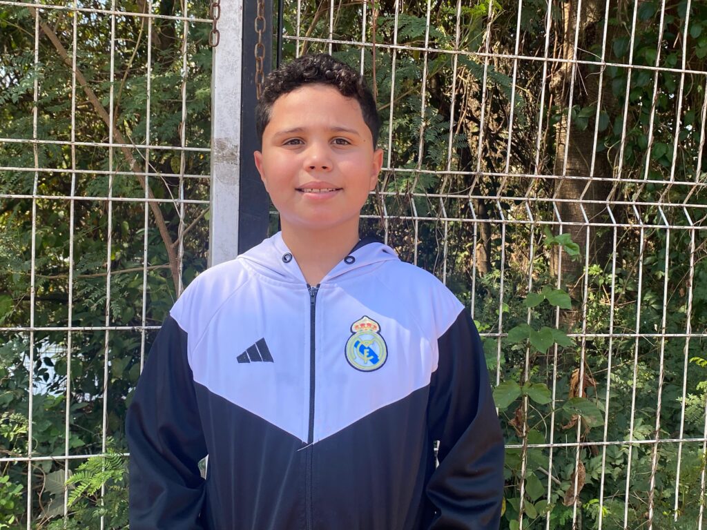 Menino de aproximadamente 8 anos sorri em frente a uma grade metálica, usando jaqueta preta e branca do time de futebol Real Madrid. Ao fundo, árvores e folhagem verde.