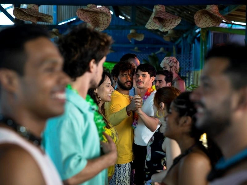 Cena de um filme brasileiro mostrando um grupo de jovens em uma festa decorada com chapéus de palha e colares havaianos. Em primeiro plano, pessoas sorriem e conversam, desfocadas. No centro da imagem, dois rapazes dançam juntos: um veste camiseta amarela e outro camiseta branca, ambos se olhando com expressão séria. Ao fundo, outros convidados completam o ambiente festivo.