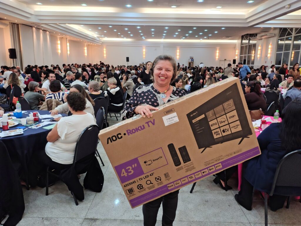 A mulher sorri para a câmera enquanto segura uma caixa de TV AOC Roku de 43 polegadas. Ela está em um salão lotado com muitas pessoas sentadas em mesas. O salão parece ser um local de evento, talvez um jantar ou uma conferência, com iluminação de teto e um palco em um lado. A caixa da TV está inclinada e a frente dela exibe uma imagem de uma TV com vários aplicativos de streaming, como Netflix, Disney+ e Amazon Prime Video. Há também um controle remoto e informações sobre a TV impressas na caixa. As pessoas nas mesas ao fundo estão conversando e interagindo, sugerindo uma atmosfera social e animada.