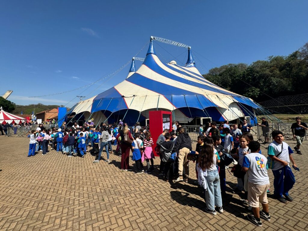 Fotografia mostra uma grande lona de circo azul e branca montada no Parque do Engenho Central em Piracicaba. Em frente à lona, dezenas de crianças e adolescentes de uniforme escolar caminham em fila e se organizam para entrar. O céu está azul e ensolarado.