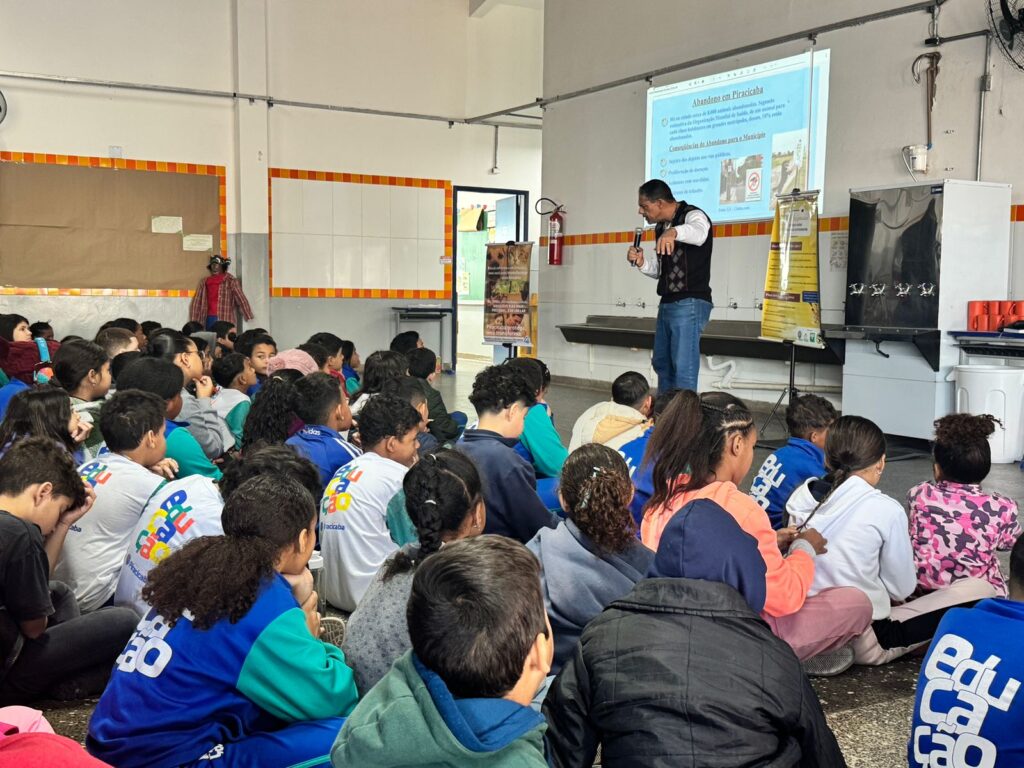 A imagem retrata uma ação educativa em uma sala de aula, onde um grupo de estudantes está sentado no chão, atento à apresentação de um homem que utiliza um microfone e aponta para uma tela de projeção. O conteúdo exibido aborda o tema “Abandono em Piracicaba” e propõe “Conscientização e Ações para a Prevenção”, sugerindo que se trata de uma palestra sobre o abandono de animais. Ao lado da tela, há um banner complementar com mais informações, reforçando o caráter informativo do evento. O ambiente é simples, com paredes brancas e detalhes em laranja, e um quadro de avisos ao fundo.