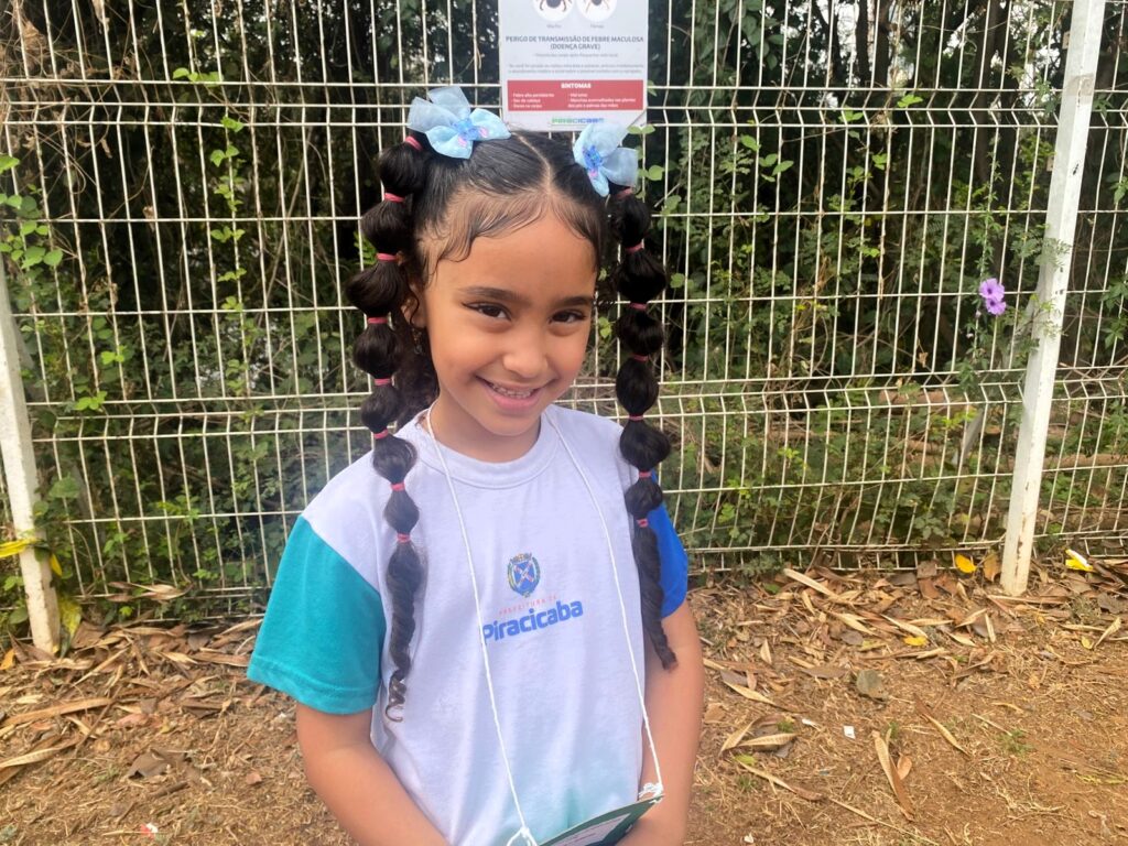 Criança com uniforme da Prefeitura de Piracicaba, cabelo preso em duas tranças volumosas presas com fitas azuis, sorri para a câmera em frente a uma cerca de arame.