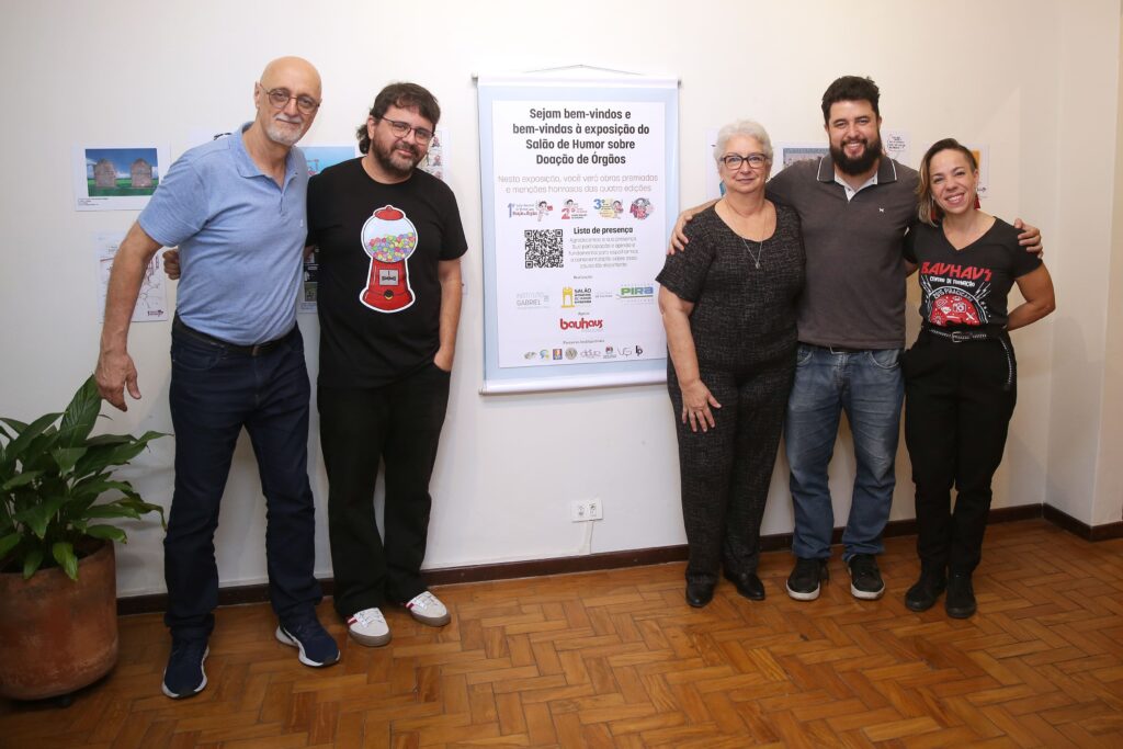 Descrição: Cinco pessoas posam sorridentes diante de um painel de boas-vindas da exposição sobre Doação de Órgãos. À esquerda, um homem de camiseta azul clara; ao lado dele, outro de barba e óculos, com camiseta preta com desenho de uma máquina de chicletes. No centro, uma senhora de cabelos brancos e óculos veste roupa preta. À direita, dois jovens: um homem de barba, com camiseta cinza, e uma mulher sorridente, com camiseta preta estampada.
