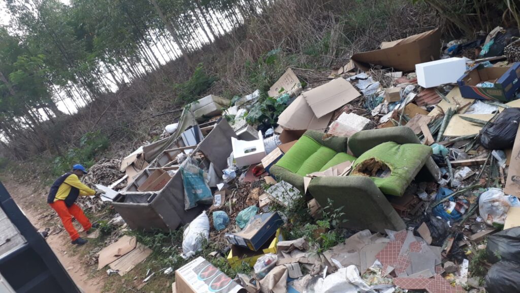 A imagem mostra uma grande quantidade de lixo acumulado em uma área externa, possivelmente uma encosta de terra com vegetação ao fundo, incluindo árvores. Há uma pessoa vestindo um colete amarelo, calça laranja e boné azul, que parece estar manuseando alguns dos detritos.O lixo é composto por diversos materiais, como caixas de papelão, pedaços de móveis (incluindo um sofá verde rasgado), sacos de lixo pretos, embalagens plásticas, pedaços de isopor e outros detritos de construção ou domésticos. A cena sugere um descarte irregular de resíduos em um local inadequado.