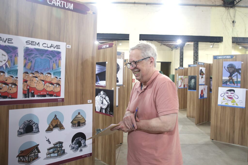 Jurado avaliando cartuns com prancheta na mão, sorrindo diante das obras.