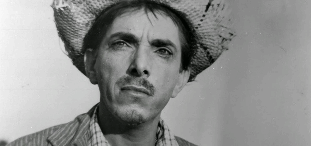 Homem de expressão séria usando chapéu de palha. Ele veste uma camisa xadrez e paletó listrado. A imagem é em preto e branco e mostra o rosto em close, com fundo neutro.