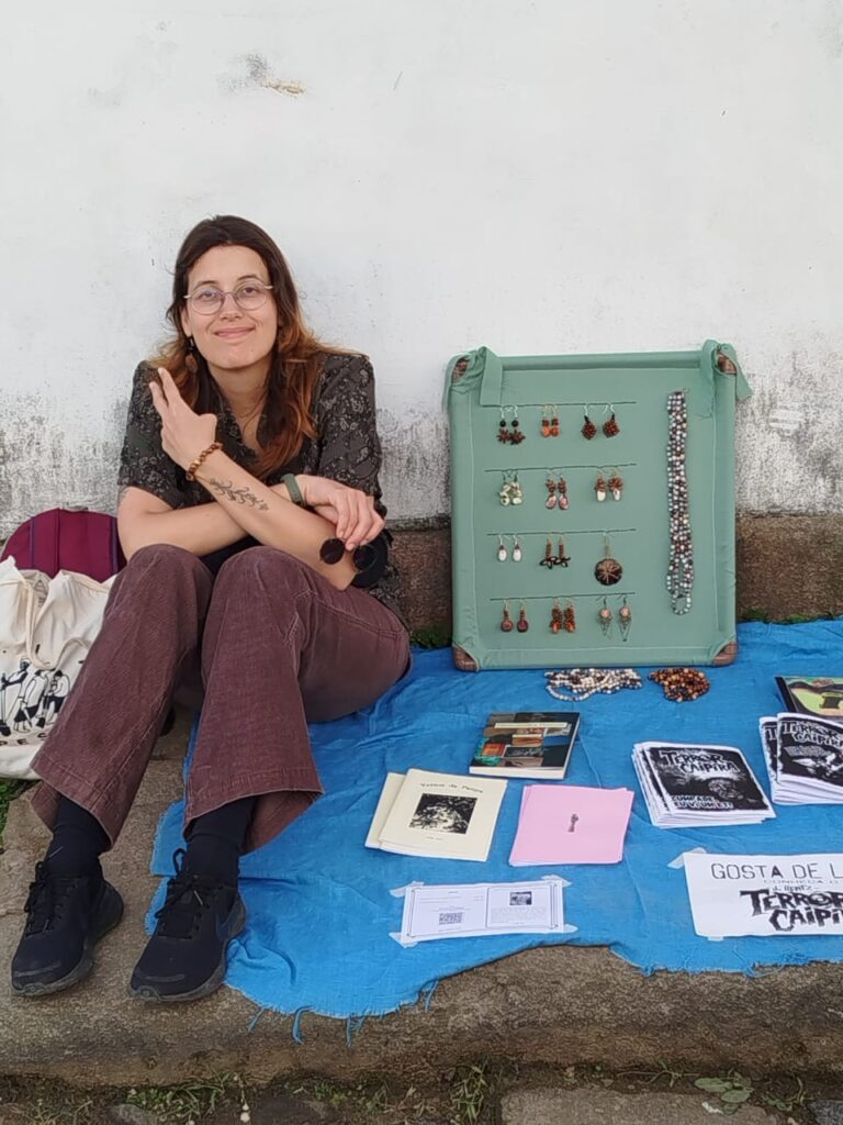 Artista Mira Mar exibe joias artesanais feitas com sementes e materiais naturais, ao lado de seus livros de poesia, compondo um espaço que valoriza sustentabilidade e arte autoral.