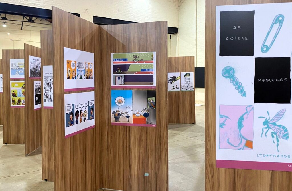 Foto de uma exposição com várias divisórias de madeira, formando corredores. Em cada painel, há cartuns coloridos com diferentes temas. Alguns têm falas de personagens, outros críticas sociais e políticas.