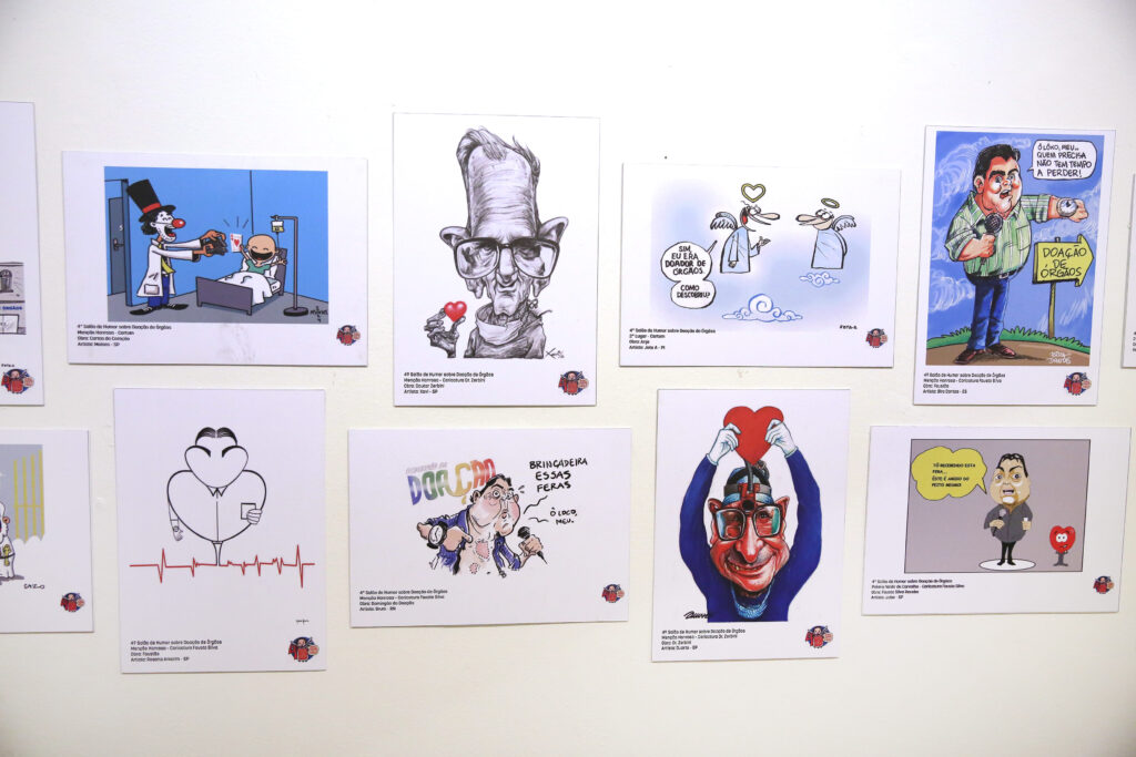 Descrição: Outra parede branca reúne oito cartuns e caricaturas coloridas sobre Doação de Órgãos. Entre eles, há desenhos de médicos, caricaturas de personalidades, uma equipe de futebol, uma figura segurando um coração e ilustrações estilizadas que destacam o tema da doação.