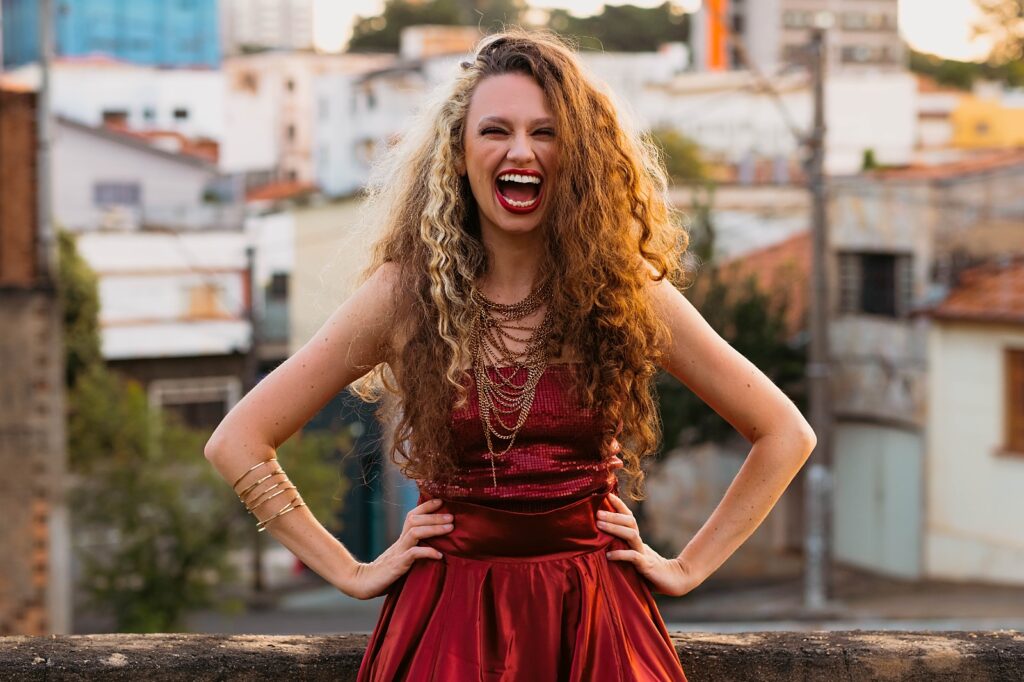 A artista está em pé ao ar livre, com as mãos na cintura, sorrindo com expressão alegre e vibrante. Ela veste um conjunto vermelho brilhante com saia rodada e colar de correntes douradas. Seus cabelos cacheados, volumosos e longos têm tons loiros e castanhos. Ao fundo, é possível ver uma paisagem urbana com casas, prédios e árvores desfocadas.