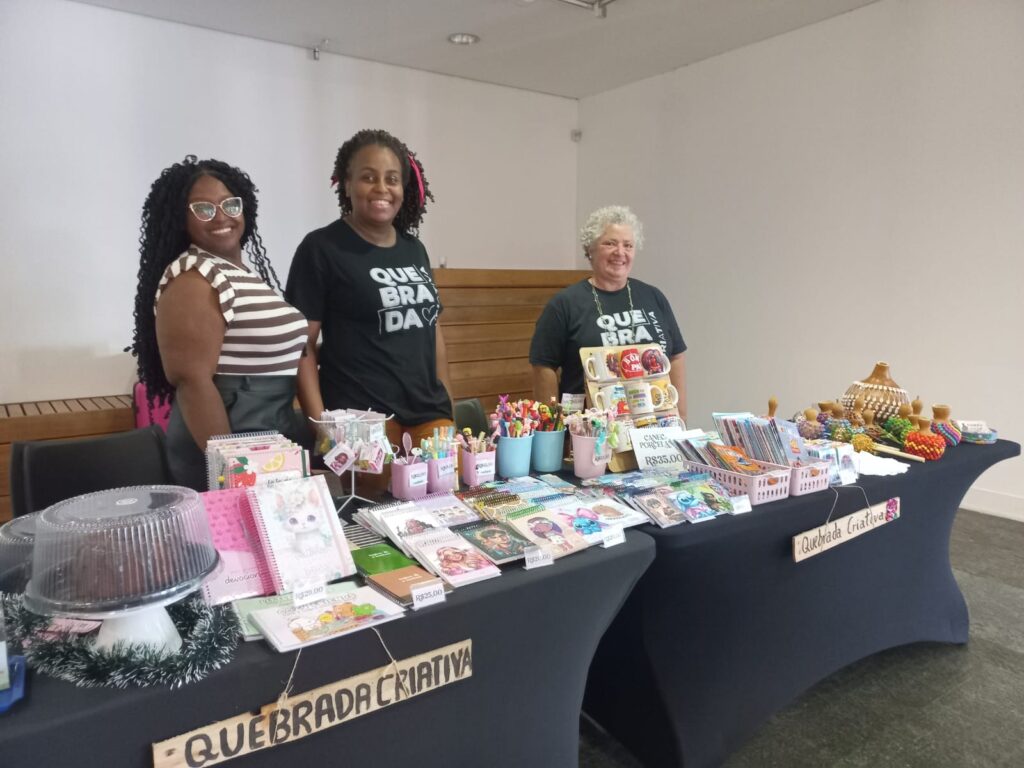 A imagem mostra três mulheres sorrindo atrás de uma mesa cheia de produtos artesanais. A mesa está coberta com uma toalha preta e expõe uma variedade de itens, incluindo cadernos, canecas, enfeites coloridos e um bolo decorado. As duas mulheres à esquerda vestem camisetas pretas com a inscrição "QUEBRADA CRIATIVA", e a mulher à direita usa uma camiseta semelhante. Há também placas com preços e nomes de produtos, como "DEVOCIONÁRIO R$20,00", "CANECAS PORCELANA R$35,00" e "QUEBRADA CRIATIVA". O ambiente parece ser uma feira de artesanato ou um evento similar.