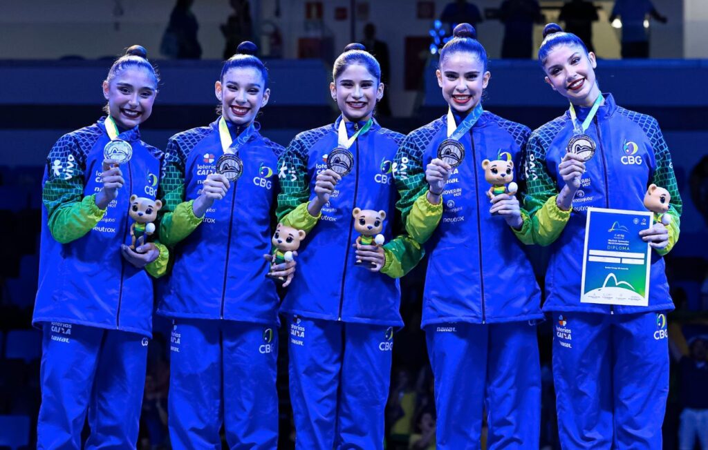 Cinco ginastas brasileiras em pé, sorrindo e exibindo medalhas e um diploma. Elas usam uniformes azuis com detalhes em verde e amarelo, e cada uma segura uma medalha de prata e um pequeno boneco de urso. A ginasta mais à direita segura um diploma. A iluminação é forte, destacando as atletas e seus prêmios em um palco ou pódio.