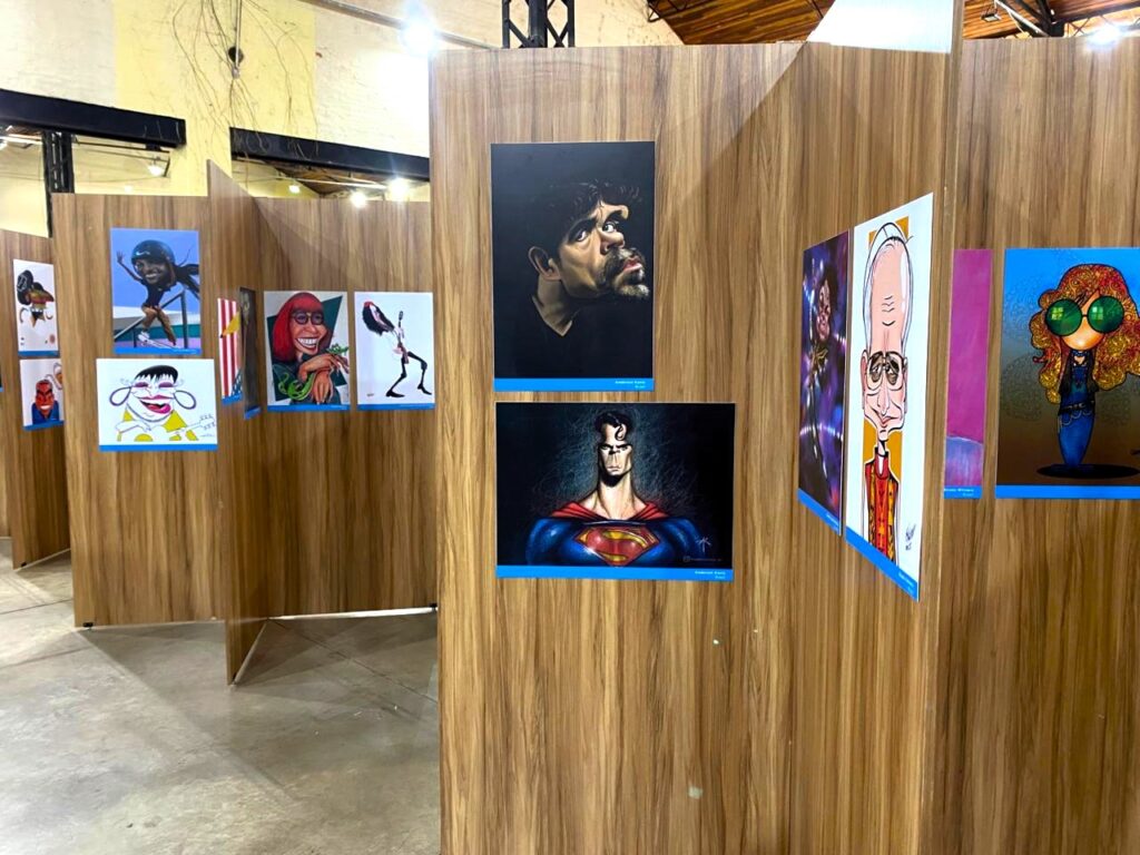 Painéis da exposição com caricaturas e desenhos. À esquerda, caricaturas exageradas de rostos e corpos de personalidades. Ao centro e à direita, imagens como o Superman com expressão séria e outros personagens desenhados em estilo cômico e artístico.