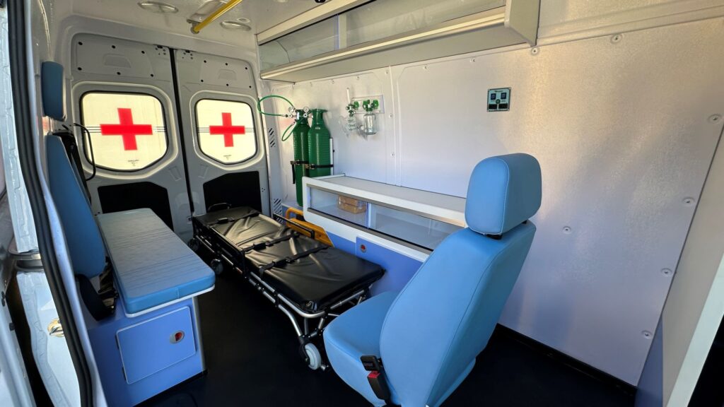 A imagem retrata o interior bem equipado de uma ambulância, destacando a estrutura organizada voltada ao atendimento médico de emergência. No centro, uma maca preta dobrável com rodas está posicionada para facilitar o transporte de pacientes, enquanto à esquerda há um banco acolchoado azul claro com compartimento inferior. Na parede do fundo, duas portas com janelas exibem cruzes vermelhas, reforçando a função do veículo. À direita da maca, dois cilindros verdes de oxigênio estão fixados com sistema de regulação e máscaras, acompanhados por uma prateleira superior para armazenamento de equipamentos. Uma cadeira adicional para o atendente médico completa o ambiente funcional e preparado para situações críticas.