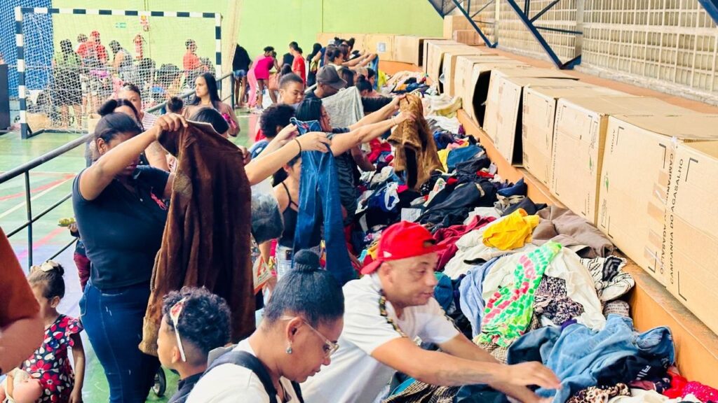A imagem mostra um grupo de pessoas dentro de um ginásio, cercado por caixas de papelão e uma grande quantidade de roupas espalhadas. As pessoas estão em pé e agachadas, selecionando e organizando as roupas. Algumas pessoas estão segurando peças de vestuário, como casacos e calças jeans. Ao fundo, há uma quadra esportiva com uma rede de gol e mais pessoas observando a cena. As caixas de papelão ao longo da parede parecem estar rotuladas com texto em português, indicando uma campanha de doação de agasalhos. A atmosfera geral sugere uma ação social ou evento de arrecadação de roupas de inverno.