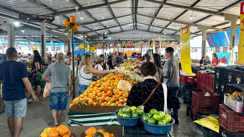 A imagem retrata um mercado coberto em pleno funcionamento, com diversos clientes circulando entre as barracas de frutas e verduras. O ambiente é vibrante e movimentado, com destaque para os montes de laranjas e limões verdes em primeiro plano, que chamam atenção pela cor e abundância. As barracas exibem placas escritas à mão com os preços dos produtos, reforçando o caráter popular e acessível do local. A estrutura metálica do teto e a disposição dos estandes indicam um espaço comunitário voltado à comercialização de alimentos frescos, evidenciando a dinâmica da economia local e o contato direto entre produtores e consumidores.