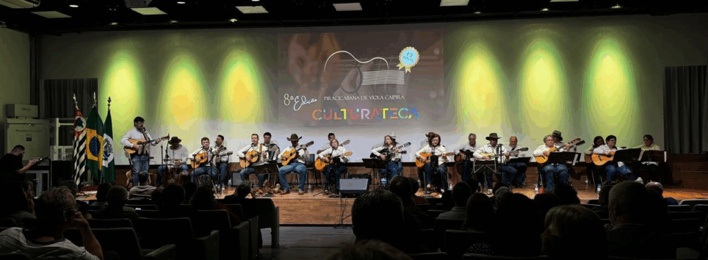 Um palco iluminado em tons de verde e amarelo recebe a Orquestra Piracicabana de Viola Caipira. Mais de 20 músicos estão sentados em fileiras, tocando viola caipira, todos vestidos com camisas claras. Ao fundo, há uma grande projeção que mostra o logotipo da orquestra e o texto “8ª edição Piracicabana de Viola Caipira – Culturateca – 19 anos”. Na frente, parte da plateia aparece assistindo atenta.