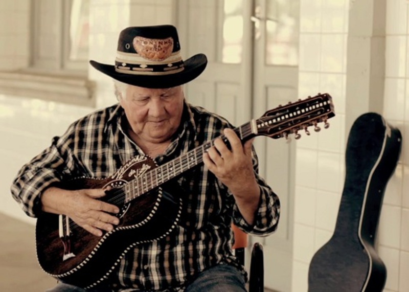 Homem idoso com chapéu e camisa xadrez toca viola caipira, com estojo do instrumento apoiado ao fundo.