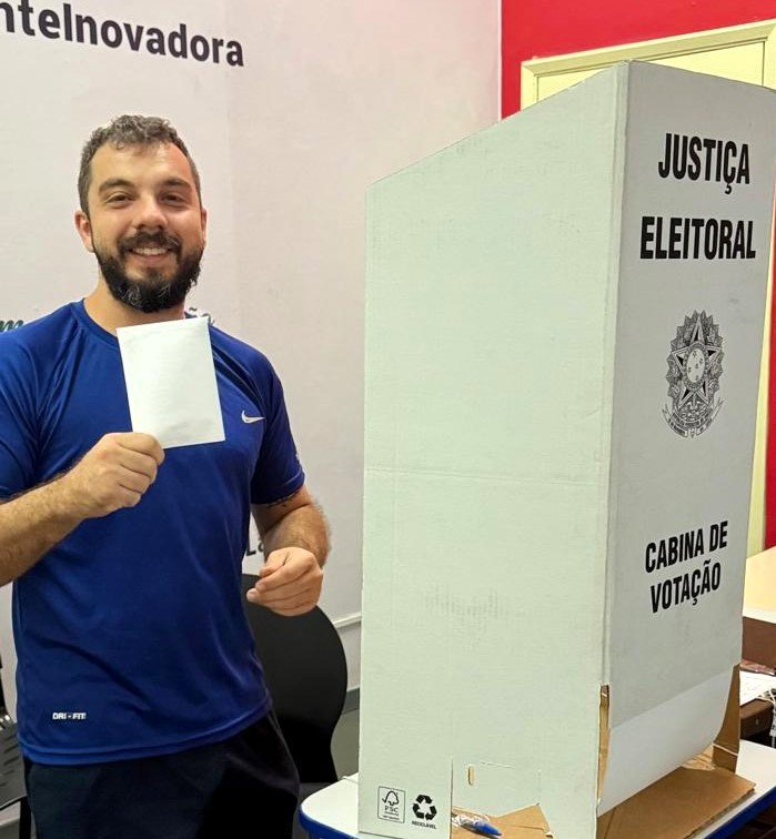 Um homem sorridente, de barba e camiseta azul, segura uma cédula de votação ao lado de uma cabine branca da Justiça Eleitoral. Ele olha para a câmera, orgulhoso por ter participado da eleição.