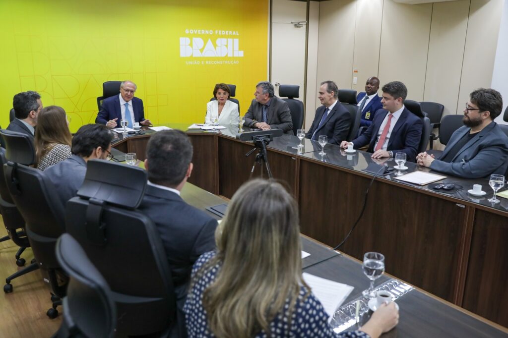 A imagem mostra uma reunião de várias pessoas sentadas em torno de uma mesa longa. No fundo, há uma parede amarela com a inscrição "GOVERNO FEDERAL BRASIL UNIÃO E RECONSTRUÇÃO". À esquerda, um homem de terno azul e gravata azul está falando e gesticulando. À sua frente, há uma mulher com uma blusa estampada. Mais à esquerda, um homem de terno escuro olha para a frente. No centro da mesa, uma mulher de terno branco está sentada, olhando para a pessoa à sua esquerda. Ao lado dela, um homem de terno cinza e gravata azul está sentado, olhando para a frente. À direita da mesa, um homem de terno azul e gravata vermelha está sentado, olhando para a frente. Ao lado dele, um homem de terno escuro com barba está sentado, com as mãos cruzadas sobre a mesa. Há copos de água e xícaras de café na mesa, juntamente com alguns papéis e um dispositivo de videoconferência. As cadeiras são pretas e giratórias. A iluminação é profissional, destacando os participantes da reunião.