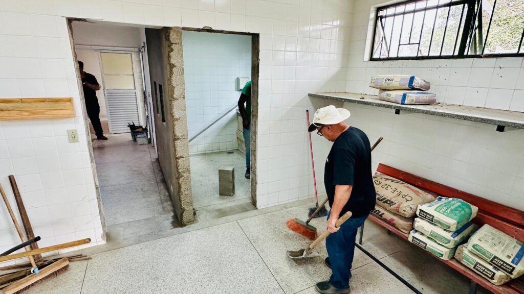 A imagem retrata uma cena de reforma ou construção dentro de um cômodo revestido por azulejos brancos. Três homens estão presentes: um em destaque segurando uma pá e uma vassoura, outro parcialmente visível por uma porta, e o terceiro ao fundo. O ambiente contém materiais de obra como sacos de cimento empilhados em prateleiras e bancos, além de ferramentas espalhadas, como níveis, pás e vassouras. Uma abertura inacabada com blocos de concreto expostos indica que o trabalho ainda está em andamento.
