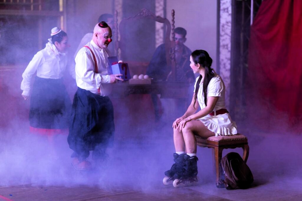 Um palhaço calvo de nariz vermelho e roupa clássica segura uma caixa com ponto de interrogação enquanto interage com uma mulher sentada em um banco, usando patins e vestido branco. Atrás deles, outra palhaça observa a cena, e há fumaça cênica cobrindo o chão. Ao fundo, um homem maquiado senta-se à frente de uma mesa com bolas de malabares. O clima é lúdico e misterioso.