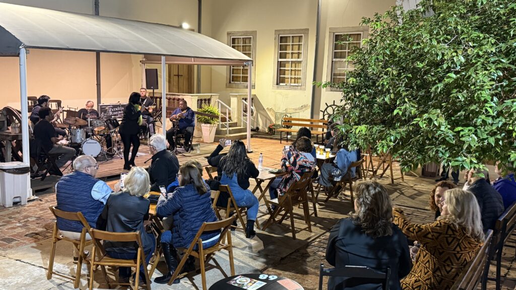 O público está sentado em mesas de madeira no quintal iluminado do Museu Prudente de Moraes, assistindo a uma apresentação musical ao vivo. No palco, há músicos tocando bateria, violão, cavaquinho e outros instrumentos, enquanto uma cantora se apresenta de pé ao centro. O cenário é acolhedor, com uma árvore frondosa à direita e iluminação amarelada que valoriza a arquitetura antiga do museu. Algumas pessoas registram o momento com o celular.