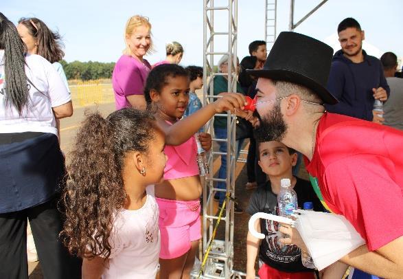 Homem fantasiado com cartola e nariz de palhaço se inclina para que uma menina coloque o nariz vermelho nele, enquanto outras crianças observam sorrindo.