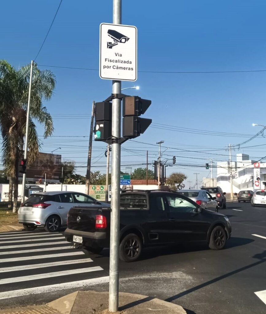 A imagem mostra uma rua em um dia ensolarado, com um céu azul claro. No centro da imagem, um poste de semáforo domina a cena, com um sinal de trânsito no topo que exibe o ícone de uma câmera e o texto "Via Fiscalizada por Câmeras". Abaixo do sinal, estão os semáforos, com as luzes verdes acesas. Na rua, há vários carros. Um carro preto, um modelo de picape, está posicionado na faixa de pedestres. Um carro prateado está logo atrás dele. Outros carros podem ser vistos mais adiante na estrada, em diferentes faixas. A rua é larga e parece ser uma via movimentada. Há também árvores e edifícios ao fundo, sugerindo um ambiente urbano. As faixas de pedestres são proeminentes na frente, com listras brancas e pretas claras.