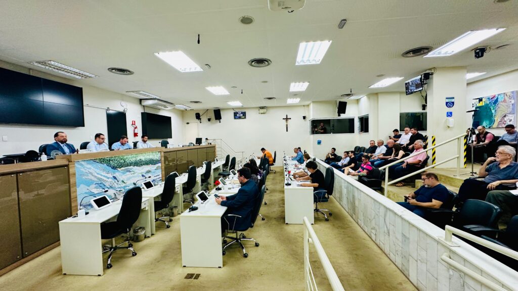 A imagem retrata uma audiência formal em uma câmara municipal ou ambiente governamental. O espaço é organizado com um corredor central ladeado por mesas equipadas com microfones e tablets, onde diversos participantes estão sentados, atentos às discussões. À esquerda, um painel elevado abriga quatro pessoas que parecem conduzir a reunião. Ao fundo, um grande mural artístico exibe uma paisagem com montanhas e rios, conferindo um toque cultural ao ambiente. À direita, o público acompanha o evento, reforçando o caráter participativo da ocasião. O ambiente é moderno e bem iluminado, com telas, câmeras de segurança e um crucifixo na parede, indicando também aspectos simbólicos ou religiosos presentes na estrutura institucional.