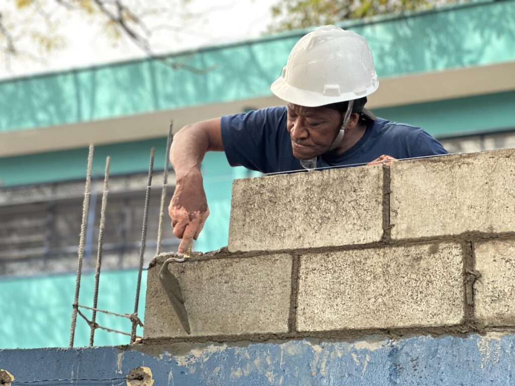A imagem retrata um trabalhador da construção civil em plena atividade, vestindo capacete branco de segurança e camisa azul escura, enquanto aplica argamassa com uma colher de pedreiro em uma parede de blocos de concreto. O cenário revela o processo de alvenaria estrutural, com vergalhões verticais visíveis que indicam reforço da estrutura. Ao fundo, observa-se um edifício com fachada em tons de turquesa e branco
