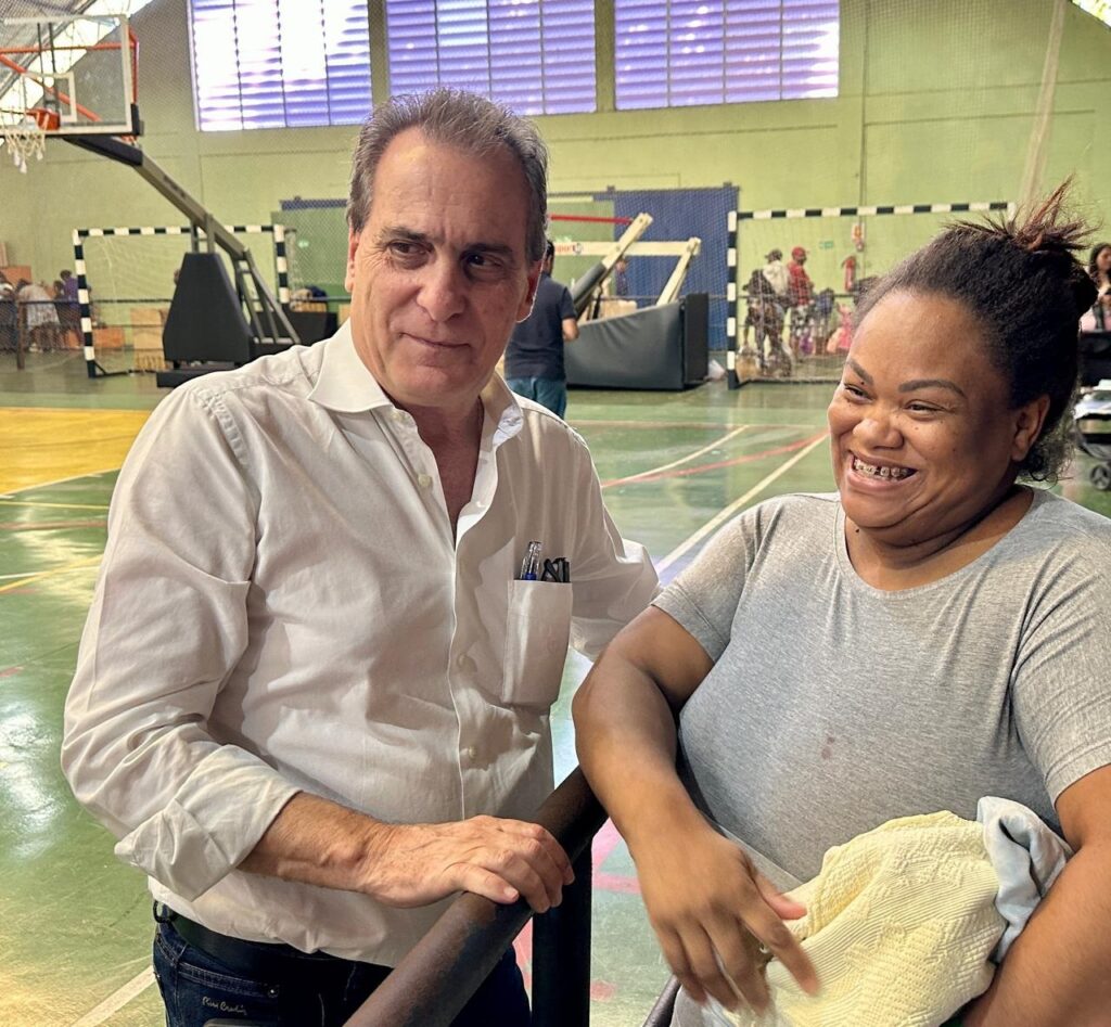 Em uma quadra esportiva, um homem de camisa social branca e um mulher de camiseta cinza estão conversando e sorrindo. O homem tem cabelos grisalhos e está com a mão apoiada em uma barra de metal. A mulher tem cabelo preso em um coque e segura um cobertor claro em seus braços. Ao fundo, outras pessoas e equipamentos esportivos podem ser vistos.