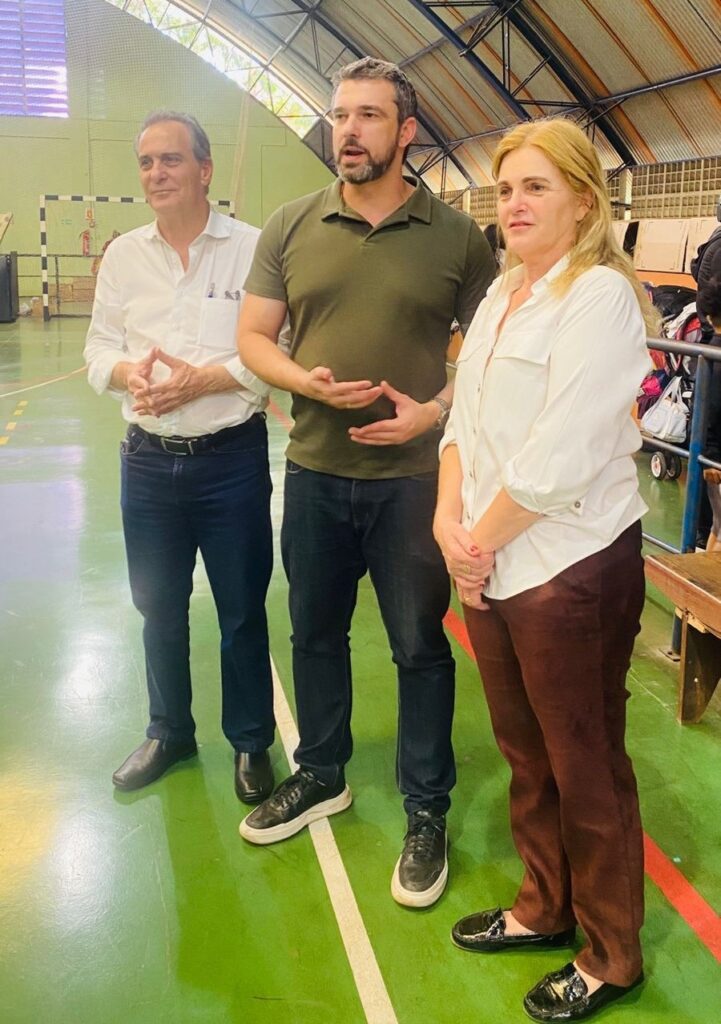 A imagem mostra três pessoas em pé em uma quadra poliesportiva. À esquerda, um homem mais velho, com cabelos grisalhos e vestindo uma camisa branca de botões e calça jeans escura, tem as mãos entrelaçadas na frente do corpo. Ao centro, um homem mais novo, com barba e cabelos escuros, usa uma camisa polo verde escura e calça jeans escura, com as mãos abertas em um gesto de fala. À direita, uma mulher loira, com uma camisa branca de botões com as mangas arregaçadas e calça marrom, olha para o lado com uma expressão séria. O fundo da imagem revela a estrutura de uma quadra, com arquibancadas e equipamentos esportivos ao fundo, sugerindo que eles estão em um evento ou visita a uma instalação esportiva. O piso da quadra é verde com marcações brancas e vermelhas.