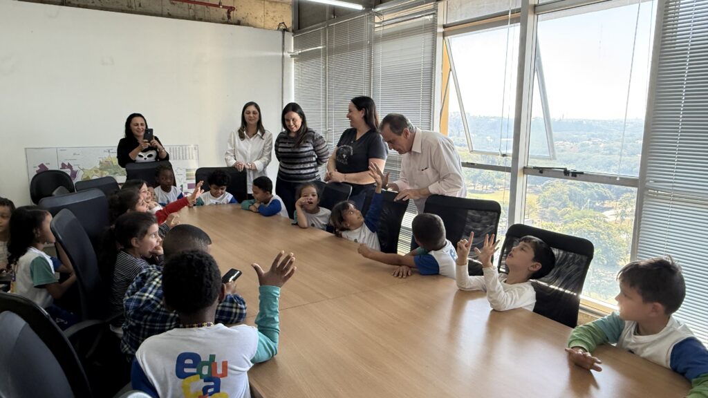 A imagem retrata um grupo de crianças sentadas ao redor de uma grande mesa de conferência, em um ambiente com janelas amplas e persianas. Algumas crianças estão com as mãos levantadas, indicando participação ativa, enquanto adultos ao fundo interagem com elas, sugerindo uma atividade educativa ou visita guiada.