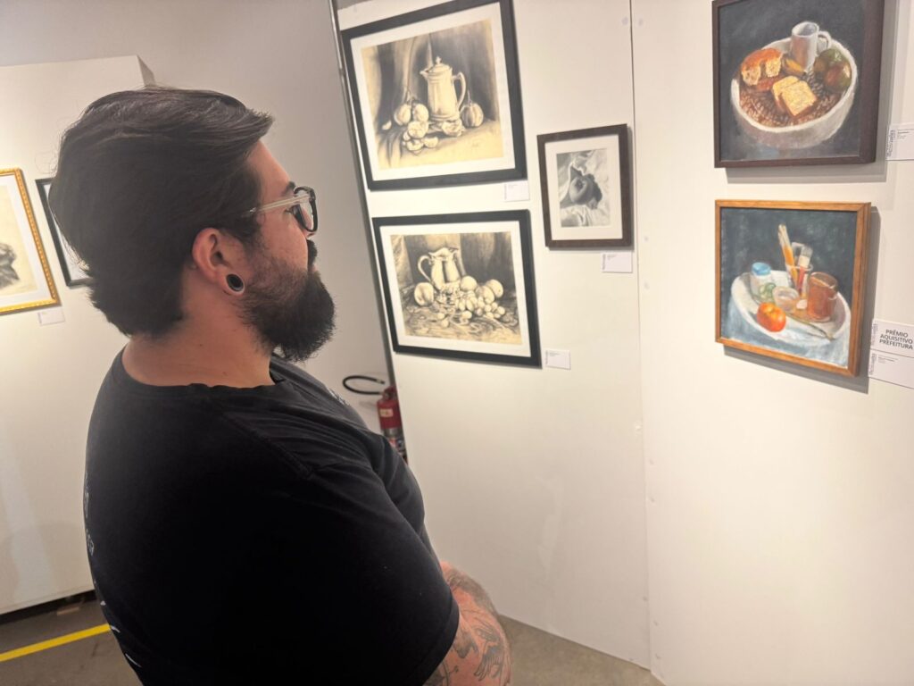 A imagem mostra um homem barbudo com óculos e alargadores nas orelhas, vestindo uma camiseta preta, olhando atentamente para uma parede branca onde estão expostas várias obras de arte. As obras são pinturas e desenhos de naturezas mortas, com destaque para frutas e objetos do cotidiano. Uma das pinturas, no canto superior direito, exibe uma tigela com pão, frutas e uma xícara. Abaixo dela, outra pintura mostra um conjunto de pincéis, potes e uma fruta sobre uma superfície branca. À esquerda, há dois desenhos em preto e branco de naturezas mortas, um com uma jarra e frutas e outro com uma jarra e legumes. Uma pequena moldura com uma imagem em preto e branco de uma figura humana está posicionada entre os desenhos. No canto inferior direito, há uma etiqueta com o texto "PRÊMIO AQUISITIVO PREFEITURA". O ambiente parece ser uma galeria de arte ou um museu.