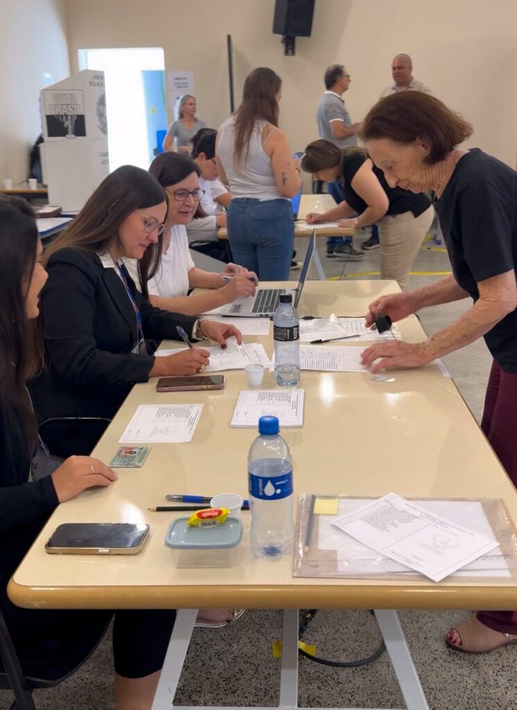 Uma senhora idosa entrega sua documentação para as mesárias em uma mesa de votação. As mulheres que trabalham como mesárias conferem os documentos e fazem registros em papéis e computadores.