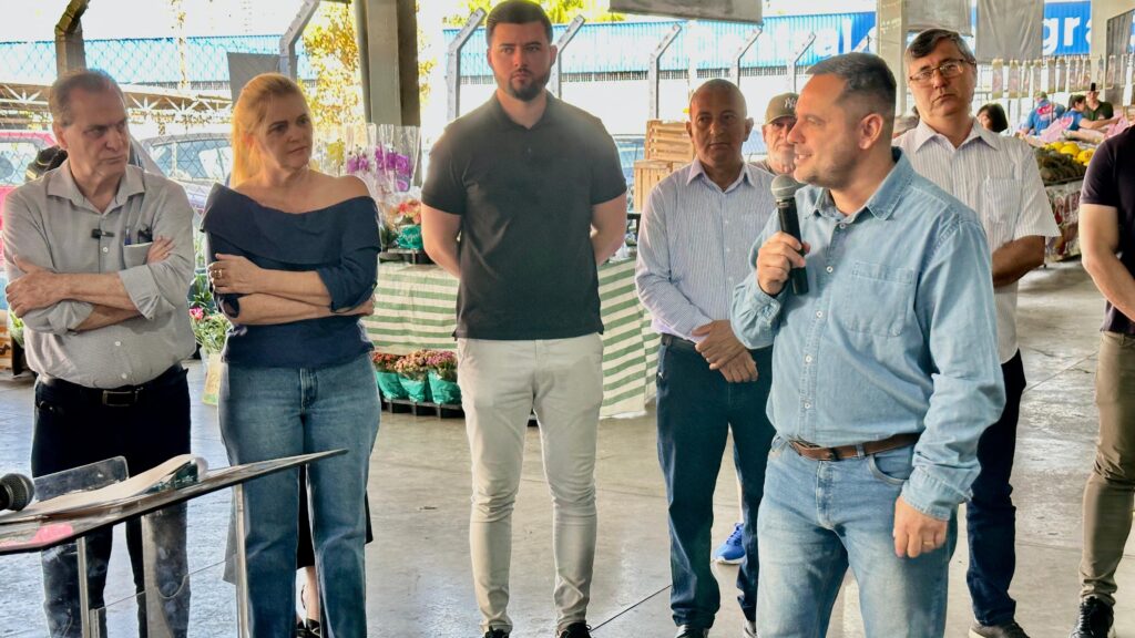 A imagem mostra um grupo de sete adultos reunidos em um espaço coberto do Varejão. Um homem à frente fala ao microfone, sugerindo que está conduzindo uma apresentação ou discurso. Ao fundo, há mesas com flores e produtos agrícolas, reforçando o ambiente de feira ou celebração local. Os participantes estão atentos e bem vestidos, com trajes que variam do casual ao social, transmitindo um clima de envolvimento e formalidade leve.