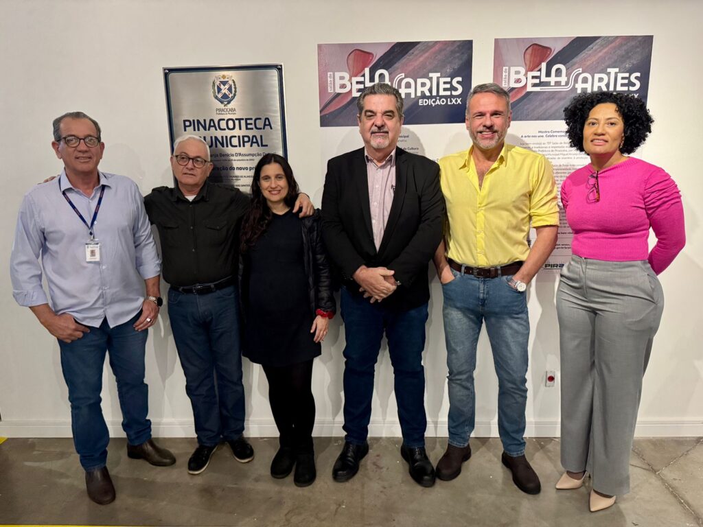 A imagem mostra um grupo de seis pessoas em frente a uma parede com um sinal e dois cartazes. Da esquerda para a direita, há um homem com camisa azul e calça jeans, um homem com camisa preta e calça jeans, uma mulher com vestido preto e jaqueta, um homem com terno preto e camisa rosa, um homem com camisa amarela e calça jeans e uma mulher com blusa rosa e calça cinza. Todos estão sorrindo e parecem estar em um evento cultural. O sinal diz "PINACOTECA MUNICIPAL" e os cartazes anunciam "BELO CARTES EDIÇÃO LXX", indicando que o evento é o 70º Salão de Belas Artes.