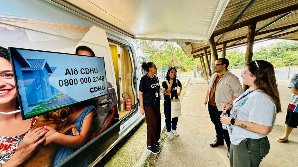 A imagem mostra uma ação comunitária promovida pela Prefeitura e pela CDHU (Companhia de Desenvolvimento Habitacional e Urbano), realizada ao lado de uma unidade móvel de atendimento. O veículo exibe um painel com o número de telefone “Alô CDHU 0800 000 2348” e uma ilustração de uma casa com uma pessoa segurando um cachorro, reforçando o vínculo com serviços habitacionais. Algumas pessoas estão reunidas em frente à van, conversando e interagindo com os atendentes, que usam camisetas com o logo da CDHU.