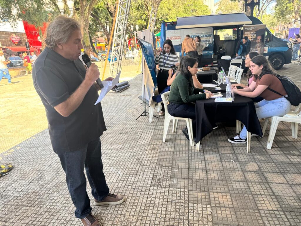 A imagem mostra um homem com cabelos grisalhos falando em um microfone, segurando papéis em sua mão. Ele está usando uma camisa polo escura e calças jeans.Ao fundo, há uma van com a inscrição "SES SENAT". Várias pessoas estão sentadas em mesas do lado de fora, algumas parecendo preencher formulários ou conversar com outras pessoas. O ambiente sugere um evento ao ar livre, possivelmente relacionado a algum serviço ou campanha educacional, dado o nome "SES SENAT" que se refere a um Serviço Social autônomo no Brasil. O piso é de cimento com um padrão quadriculado.