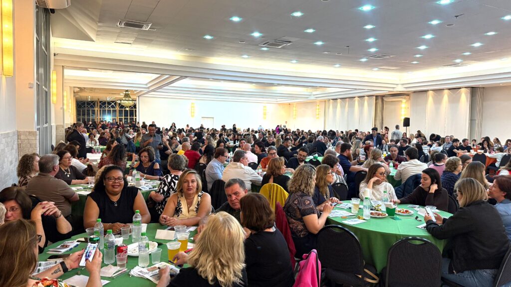 A imagem mostra um grande salão de eventos com muitas pessoas sentadas em mesas redondas, todas cobertas com toalhas verdes. O ambiente é iluminado por luzes de teto e algumas luminárias nas paredes. As pessoas estão vestidas de forma casual e parecem estar participando de um evento, possivelmente um jantar ou uma conferência, pois há garrafas de água e copos nas mesas. Em algumas mesas, há papéis e materiais que sugerem alguma atividade ou apresentação. O salão é amplo, com um teto alto e janelas grandes em uma das paredes, que revelam uma vista noturna do exterior com luzes acesas. Há também um lustre grande e decorativo no fundo do salão.