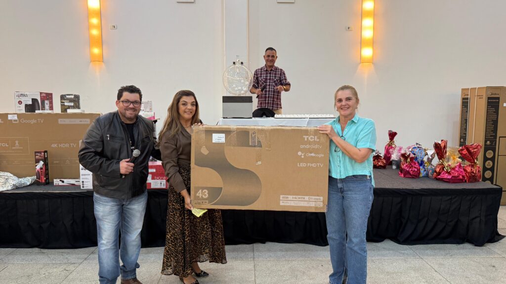 A imagem mostra um evento de sorteio. No centro, três pessoas posam para uma foto segurando uma caixa grande de televisão TCL de 43 polegadas. Uma mulher à esquerda e outra à direita seguram a caixa, enquanto um homem no centro segura um microfone. Ao fundo, um homem com uma camisa xadrez está perto de uma urna de sorteio com bolas dentro. Há várias caixas de outros produtos eletrônicos e presentes embrulhados em cima de mesas cobertas com toalhas pretas. A iluminação do local é feita por luminárias de parede com luzes amarelas.