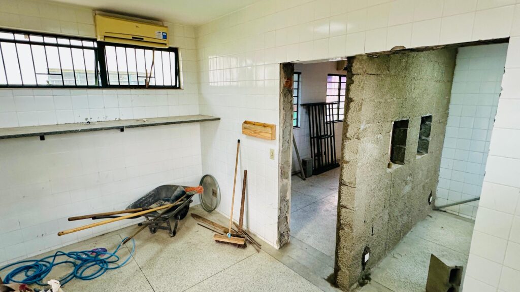 A imagem mostra um ambiente interno em processo de reforma, com paredes revestidas por azulejos brancos e piso de concreto ou granilite. Há uma parede parcialmente demolida, revelando blocos de concreto e vergalhões, o que indica que a obra está em andamento. Espalhados pelo espaço, vemos ferramentas como carrinho de mão, pá, vassoura, rastelo e um rolo de corda azul, além de uma prateleira fixada na parede. Acima dela, há uma janela com grades e um aparelho de ar-condicionado instalado. Através da abertura na parede, é possível ver outro cômodo semelhante, também com janelas gradeadas.