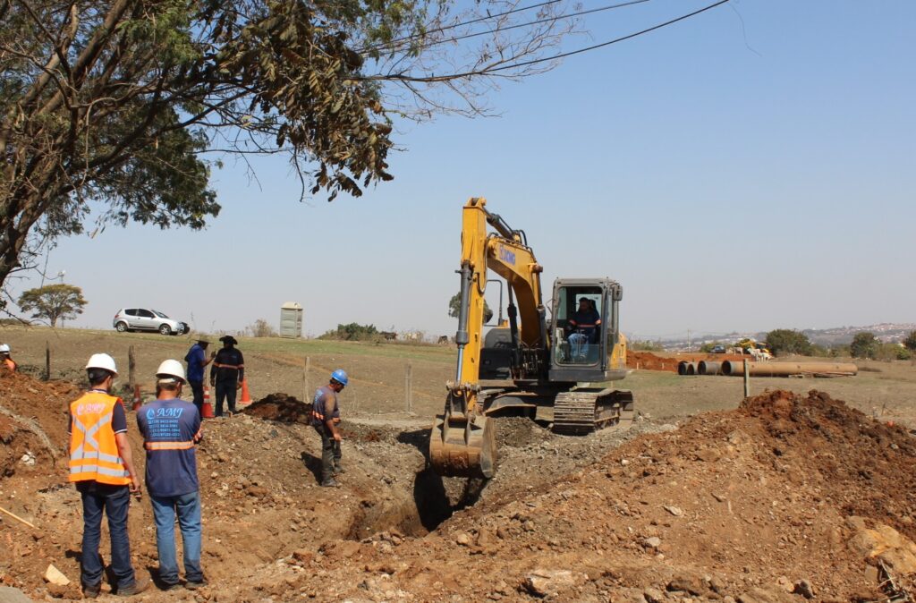 A imagem mostra um canteiro de obras em um dia ensolarado. No centro da imagem, uma escavadeira amarela está em operação, com um operador dentro da cabine. A escavadeira está cavando uma vala no solo. Ao redor da escavadeira, há vários trabalhadores da construção civil. Alguns usam capacetes brancos e coletes laranja de alta visibilidade, enquanto outros usam capacetes azuis e roupas de trabalho. Eles parecem estar supervisionando o trabalho ou realizando outras tarefas no local. No fundo, é possível ver uma área com algumas árvores, um carro prateado estacionado, um banheiro químico portátil e algumas peças de tubulação de grande diâmetro. O terreno é irregular, com montes de terra e uma vala recém-escavada. O céu está limpo e azul, indicando bom tempo. A cena geral transmite uma atmosfera de trabalho ativo e progresso na construção.
