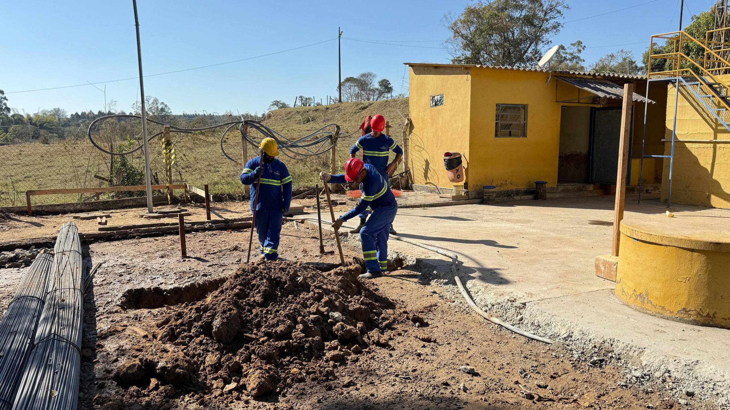 Na imagem, três trabalhadores estão em um canteiro de obras sob um céu azul claro. Eles vestem macacões azuis com detalhes em verde e amarelo, e dois deles usam capacetes vermelhos, enquanto um usa um capacete amarelo. À esquerda, há um monte de vergalhões de metal amarrados. O solo à frente é uma mistura de terra solta e pedras, com uma vala recém-cavada no centro. No centro da imagem, dois trabalhadores estão ajoelhados ou agachados, usando ferramentas para cavar ou trabalhar na terra. Um deles está usando uma pá e o outro parece estar usando uma ferramenta de mão para cavar. Ao fundo, há um edifício amarelo com um telhado cinza e uma janela pequena. Ao lado do edifício, há uma escada azul e amarela que leva a uma plataforma elevada. O local é cercado por uma cerca de arame e vegetação ao fundo, sugerindo um ambiente rural ou suburbano. A atmosfera geral é de trabalho ao ar livre sob luz solar forte.