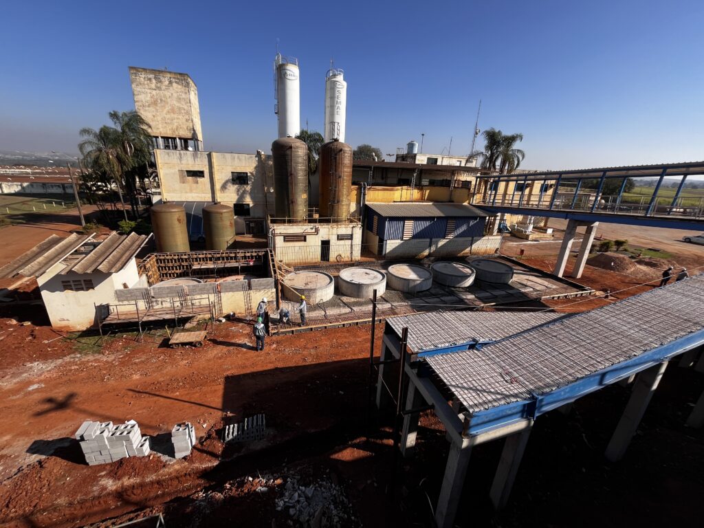 A imagem mostra uma instalação industrial com várias estruturas. Há dois tanques cilíndricos altos e brancos com a inscrição "S AMAE" no topo. Ao lado deles, há dois tanques maiores e mais escuros, com aparência de metal enferrujado. Em primeiro plano, há uma área de construção com terra vermelha e materiais de construção, como blocos de concreto empilhados. Várias pessoas, vestindo capacetes e coletes de segurança, estão trabalhando na área. Há estruturas de andaimes e formas de concreto sendo preparadas. No centro da imagem, há uma série de tanques circulares de concreto, alguns dos quais parecem estar em construção ou em processo de acabamento. Ao fundo, há um prédio de alvenaria com uma torre inclinada e alta, além de outras construções menores. Uma passarela elevada com estrutura azul e piso quadriculado atravessa a cena, conectando diferentes partes da instalação. O céu está limpo e azul, indicando um dia ensolarado.