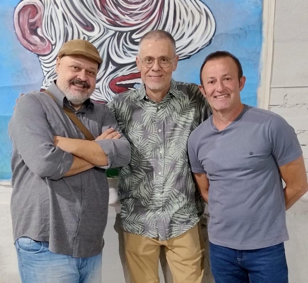 Descrição: Três homens estão em pé sorrindo para a câmera. À esquerda, um homem usa boina marrom, camisa cinza e calça jeans. No centro, um homem de óculos, barba e cabelo grisalho, veste camisa estampada com folhas verdes e calça bege. À direita, outro homem veste camiseta cinza e calça jeans. Ao fundo, uma obra de arte com traços expressivos em azul, branco e vermelho.