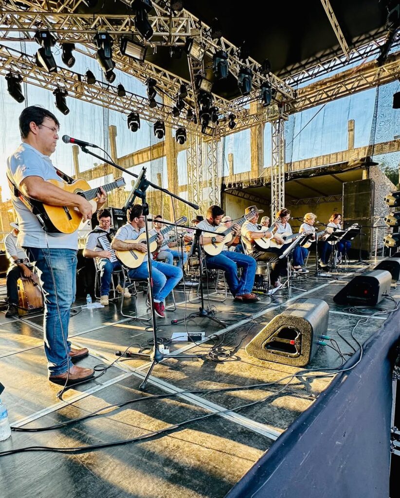 Vários músicos estão alinhados no palco, todos com violas caipiras nas mãos. Em primeiro plano, um homem toca próximo ao microfone, usando jeans e camisa clara. Ao fundo, nota-se uma estrutura de palco metálica com refletores e o pôr do sol iluminando os músicos.
